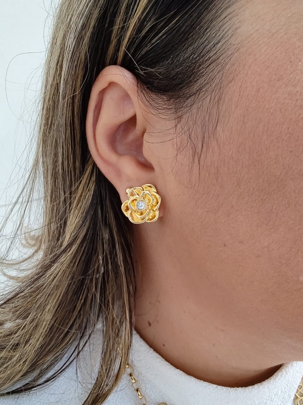 Brinco dourado em forma de flor com cristal ao centro, usado por uma mulher.