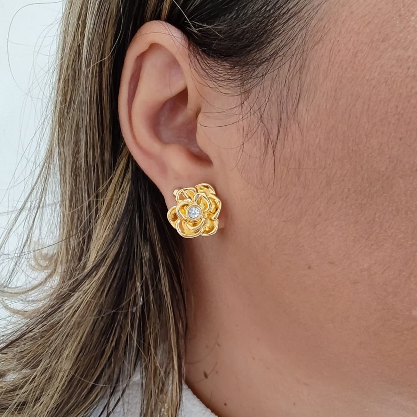Brinco dourado em forma de flor com cristal ao centro, usado por uma mulher.