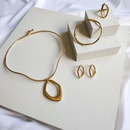 Conjunto de colar, brincos, anel e bracelete dourados com formas assimétricas sobre fundo claro.