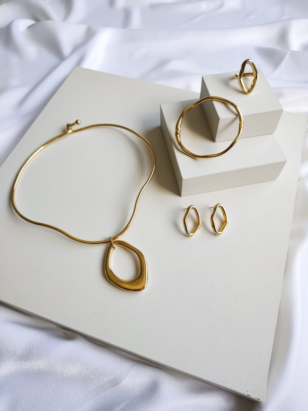 Conjunto de colar, brincos, anel e bracelete dourados com formas assimétricas sobre fundo claro.