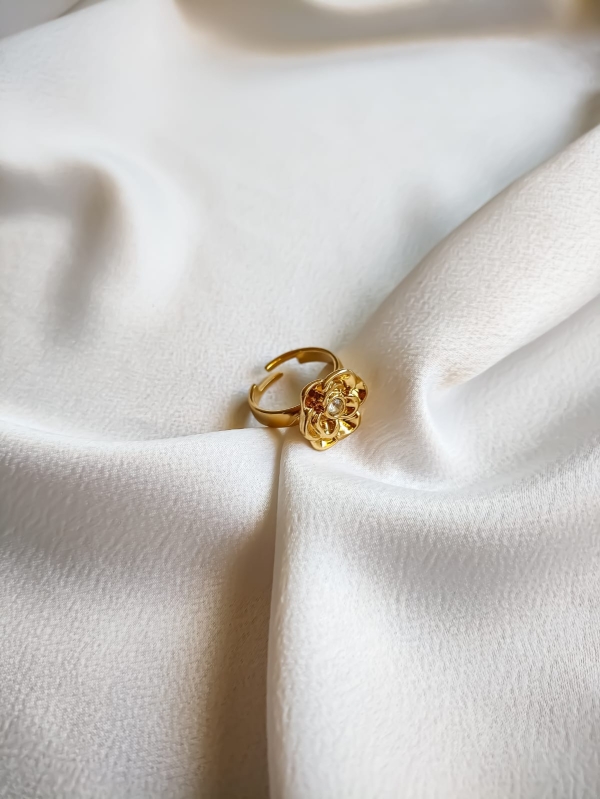 Anel dourado com flor detalhada e pedra de zircônia no centro, apoiado sobre tecido branco