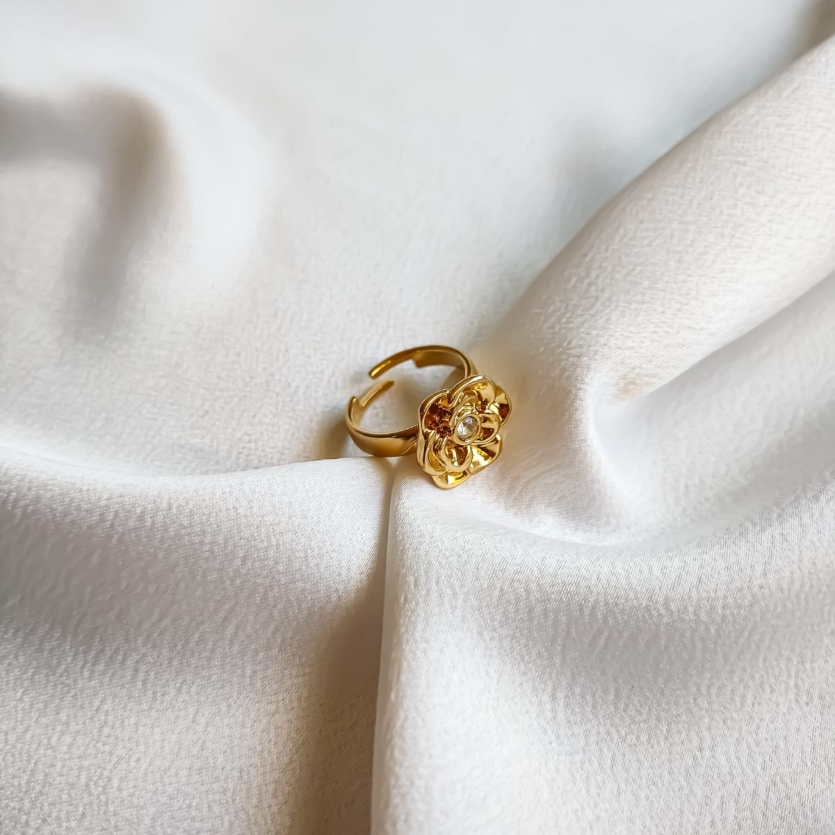 Anel dourado com flor detalhada e pedra de zircônia no centro, apoiado sobre tecido branco