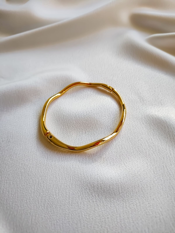 Pulseira dourada com formato ondulado e acabamento brilhante sobre tecido branco.