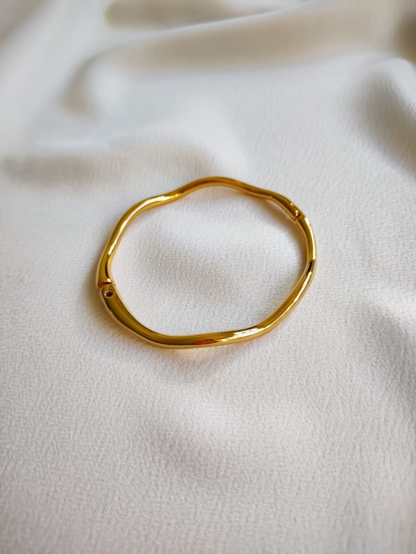 Pulseira dourada com formato ondulado e acabamento brilhante sobre tecido branco.