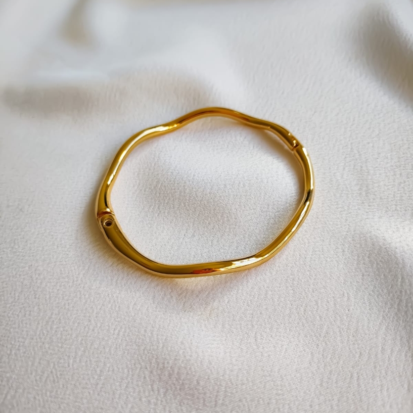 Pulseira dourada com formato ondulado e acabamento brilhante sobre tecido branco.