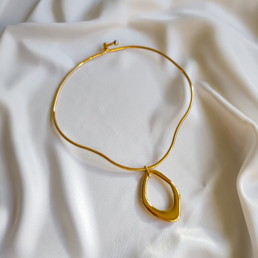 Colar rígido dourado com pingente de formato orgânico, fotografado sobre tecido branco.