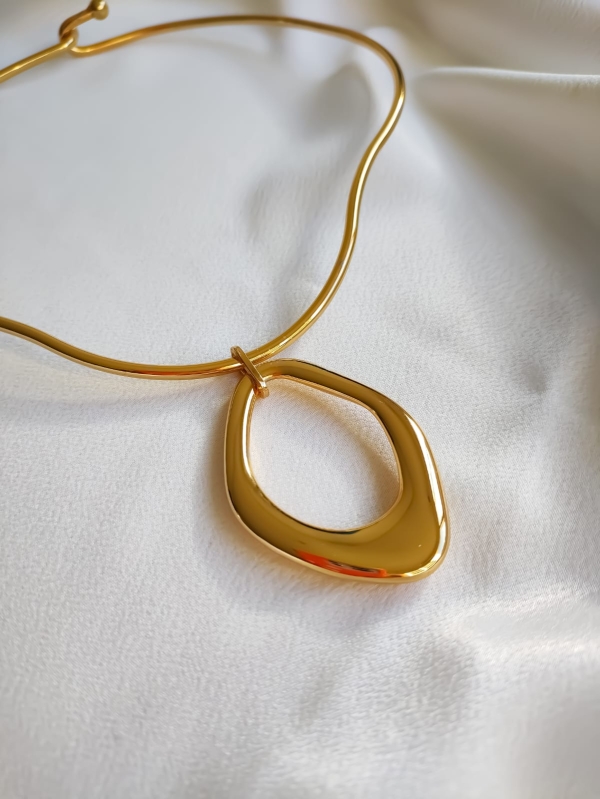 Texto alternativo (alt): Colar rígido dourado com pingente de formato orgânico, fotografado sobre tecido branco.