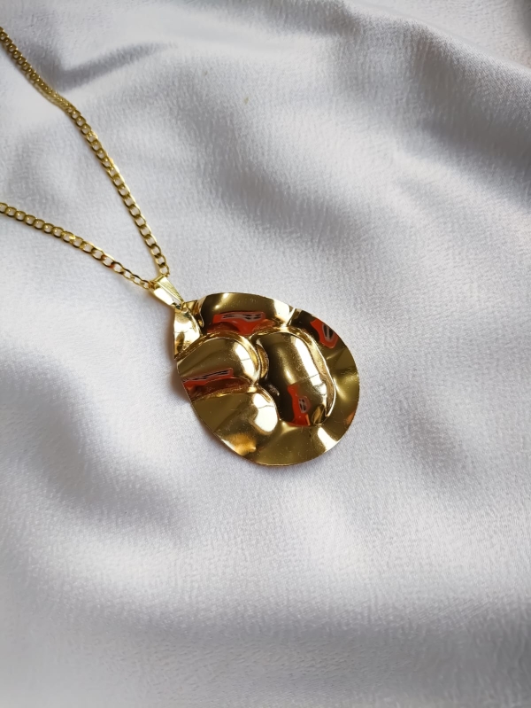 Colar dourado com pingente em formato de gota de linhas orgânicas, sobre fundo de tecido branco.