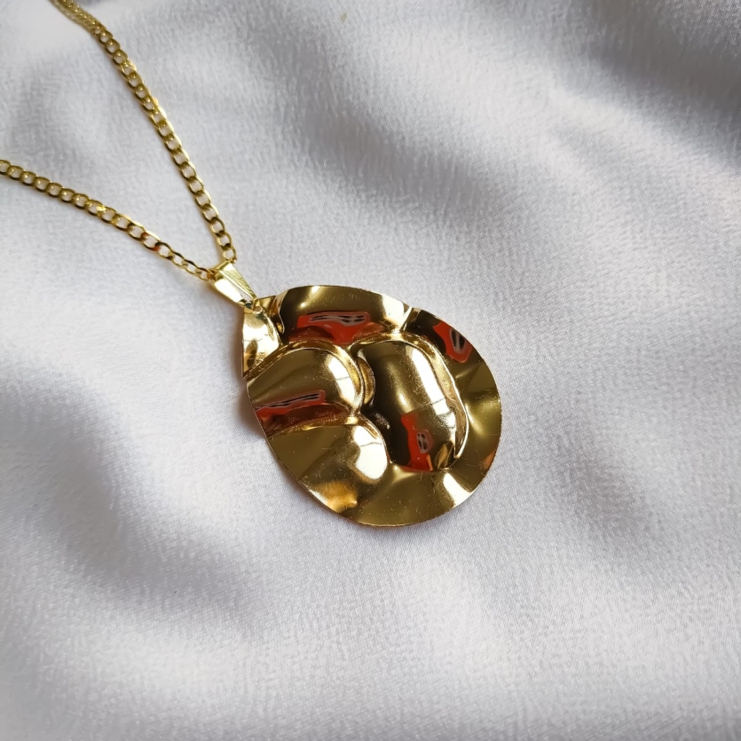 Colar dourado com pingente em formato de gota de linhas orgânicas, sobre fundo de tecido branco.