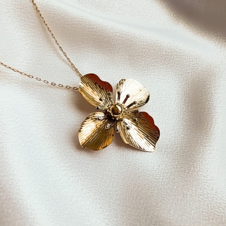 Colar dourado com pingente de flor texturizada sobre tecido branco.
