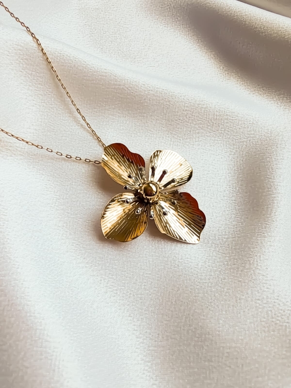 Colar dourado com pingente de flor texturizada sobre tecido branco.