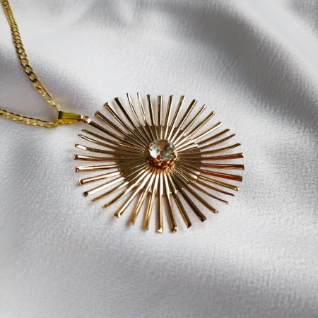 Colar dourado com pingente inspirado em raio de sol e detalhe de cristal no centro, fotografado sobre tecido branco.