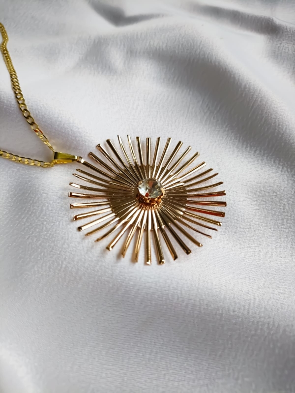 Colar dourado com pingente inspirado em raio de sol e detalhe de cristal no centro, fotografado sobre tecido branco.