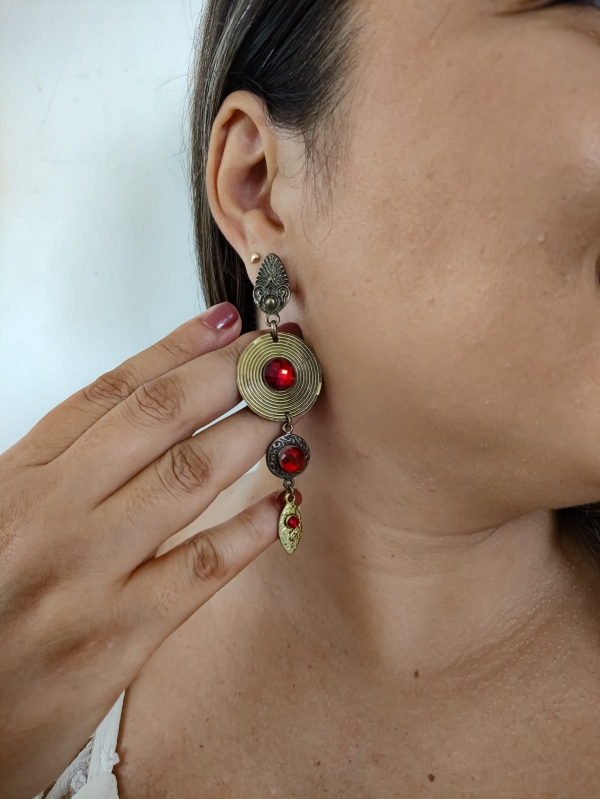 Brinco longo em estilo boho com detalhes dourados e cristais vermelhos, usado na orelha de uma mulher.