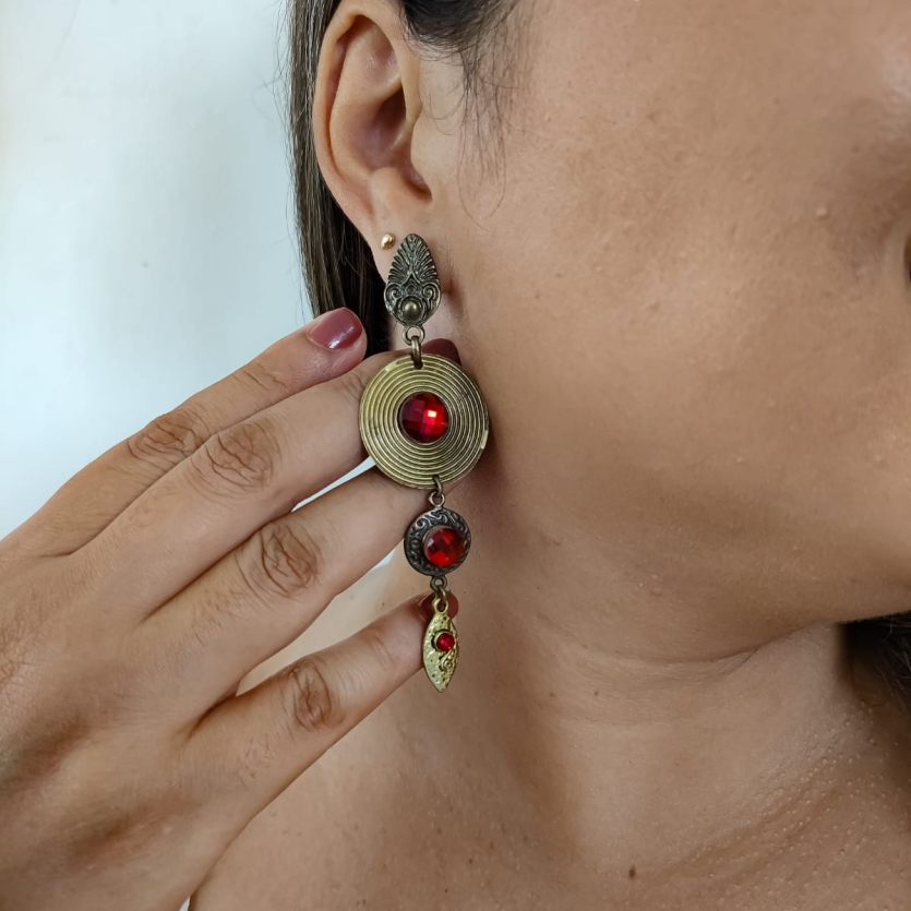 Brinco longo em estilo boho com detalhes dourados e cristais vermelhos, usado na orelha de uma mulher.