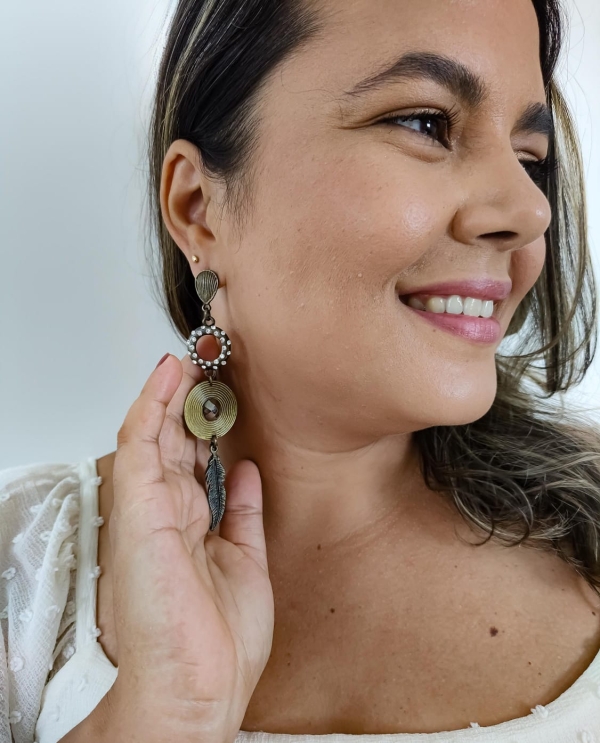 Brinco longo estilo boho com aro adornado por pérolas, disco dourado e pingente de pena, usado por uma mulher sorridente.