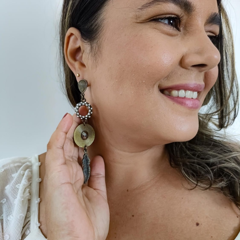 Brinco longo estilo boho com aro adornado por pérolas, disco dourado e pingente de pena, usado por uma mulher sorridente.