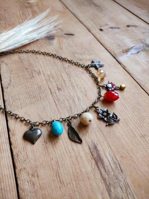 Colar boho com corrente de metal envelhecido e pingentes variados coloridos em formato de coração, flor, pedras e borboleta, sobre fundo de madeira rústica.