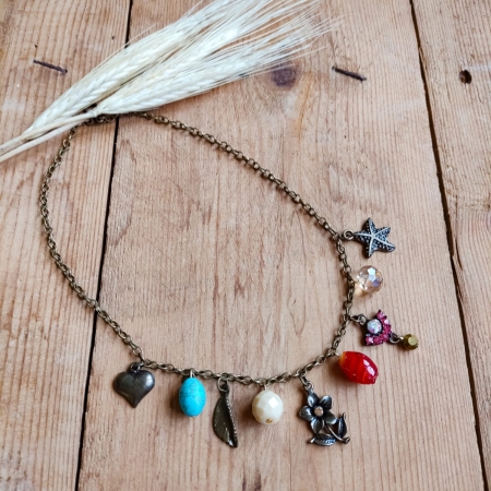 Colar boho com corrente de metal envelhecido e pingentes variados coloridos em formato de coração, flor, pedras e borboleta, sobre fundo de madeira rústica.