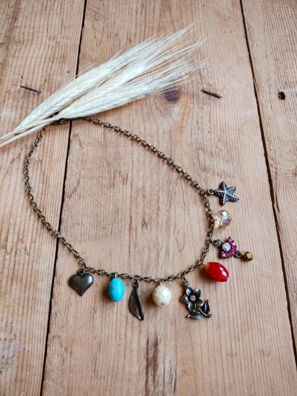 Colar boho com corrente de metal envelhecido e pingentes variados coloridos em formato de coração, flor, pedras e borboleta, sobre fundo de madeira rústica.