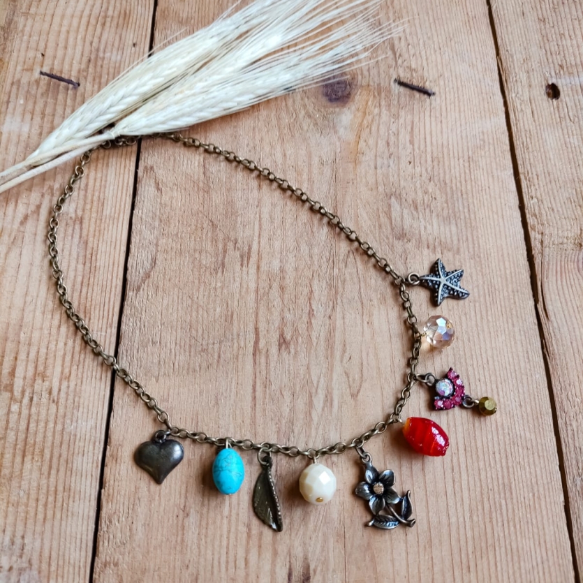 Colar boho com corrente de metal envelhecido e pingentes variados coloridos em formato de coração, flor, pedras e borboleta, sobre fundo de madeira rústica.