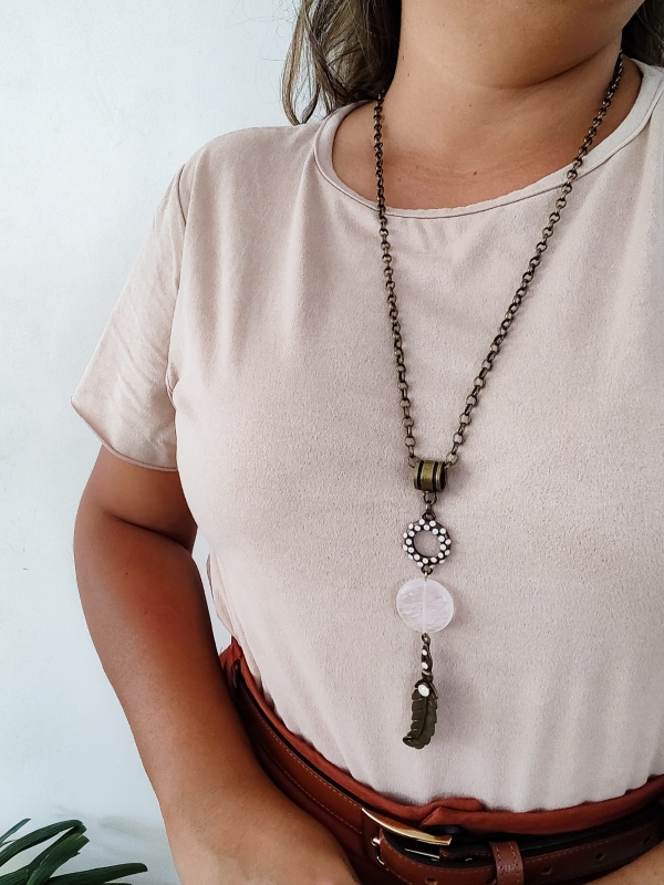 Colar com corrente de metal envelhecido, pedra branca central e pingente de folha com detalhes em strass, usado sobre blusa clara.
