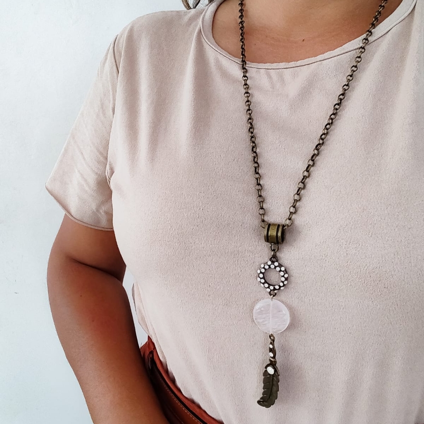 Colar com corrente de metal envelhecido, pedra branca central e pingente de folha com detalhes em strass, usado sobre blusa clara.