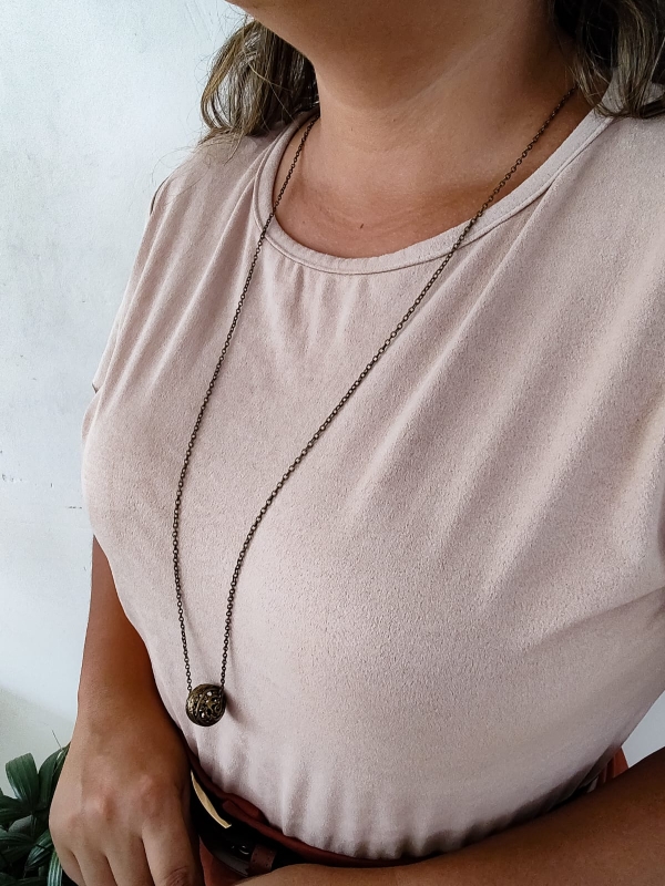 Colar longo com corrente fina e pingente em formato de esfera em metal envelhecido, usado sobre blusa clara.