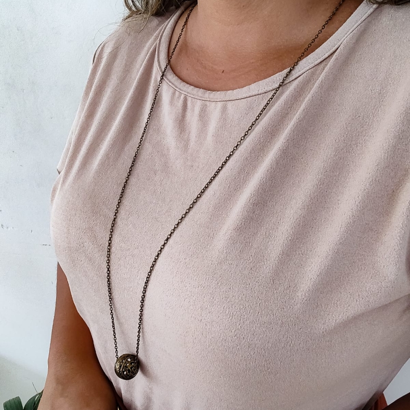 Colar longo com corrente fina e pingente em formato de esfera em metal envelhecido, usado sobre blusa clara.
