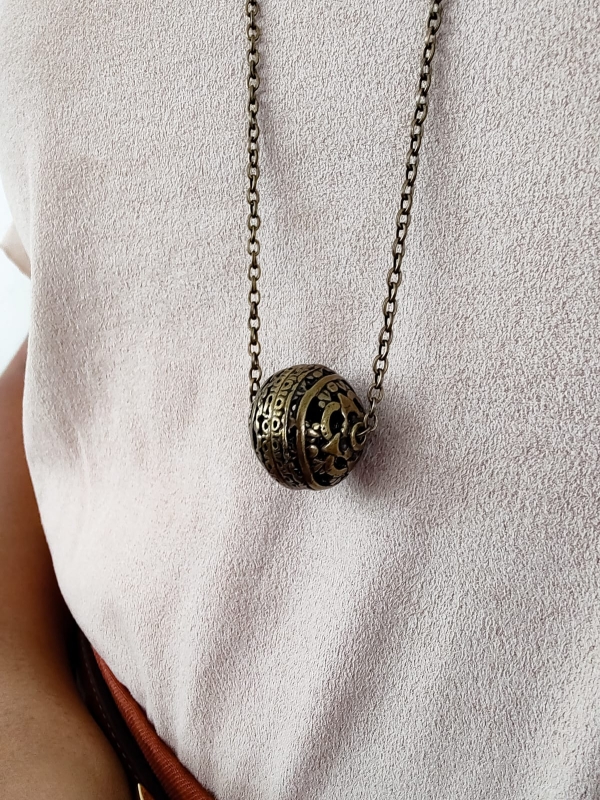 Colar longo com corrente fina e pingente em formato de esfera em metal envelhecido, usado sobre blusa clara.