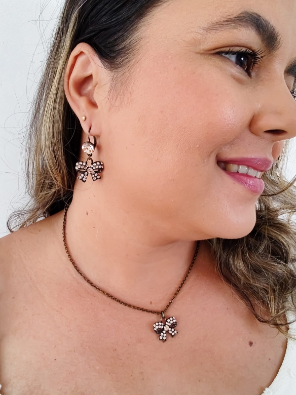 Conjunto com colar e brinco de laço em metal envelhecido com cristais, usado por mulher sorridente.
