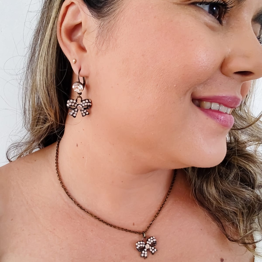 Conjunto com colar e brinco de laço em metal envelhecido com cristais, usado por mulher sorridente.