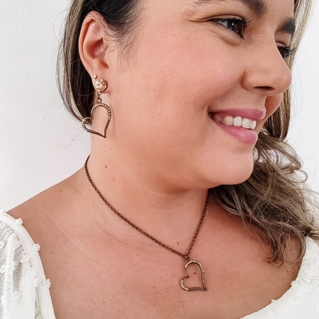 Mulher usando conjunto de colar e brinco em formato de coração vazado com cristais em metal envelhecido.