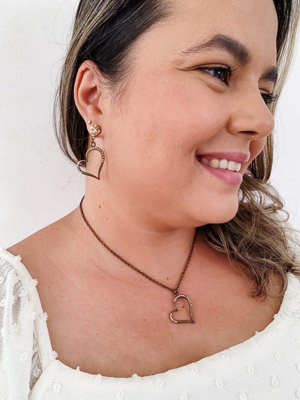 Mulher usando conjunto de colar e brinco em formato de coração vazado com cristais em metal envelhecido.