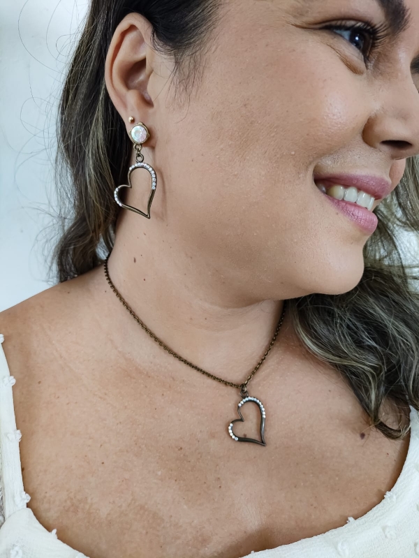 Mulher usando conjunto de colar e brinco em formato de coração vazado com cristais em metal envelhecido.