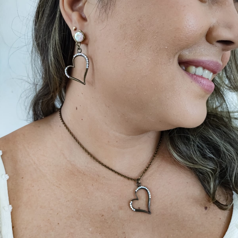 Mulher usando conjunto de colar e brinco em formato de coração vazado com cristais em metal envelhecido.