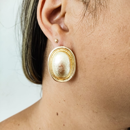 Brinco dourado oval com textura fosca usado por mulher.