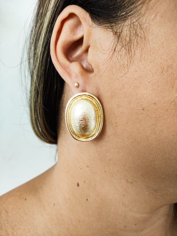 Brinco dourado oval com textura fosca usado por mulher.