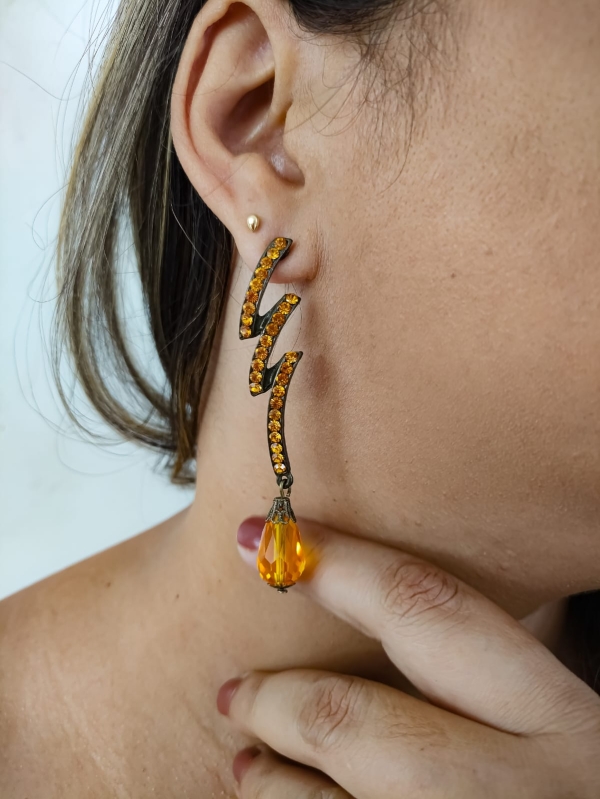 Brinco longo dourado com pedras laranja em zigue-zague e gota âmbar pendente usado por mulher.