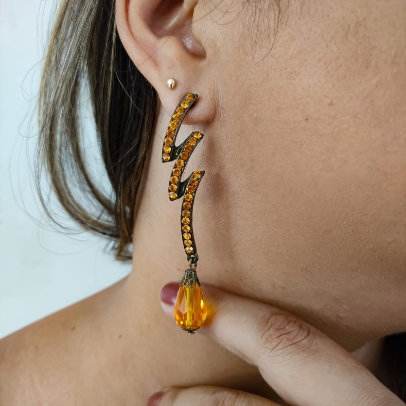 Brinco longo dourado com pedras laranja em zigue-zague e gota âmbar pendente usado por mulher.