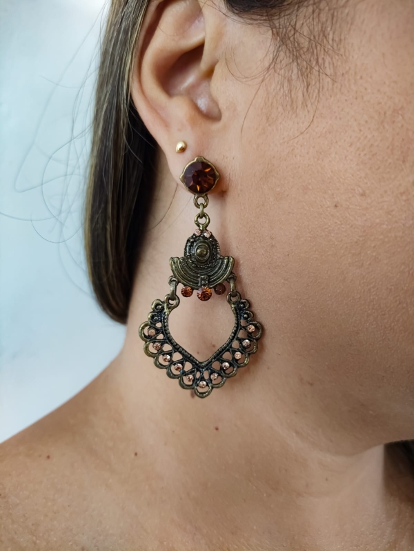 Brinco boho em tom bronze com detalhe em formato de coração vazado e cristais âmbar, usado na orelha feminina.