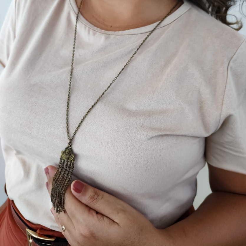 Colar longo com pingente bronze e correntes finas estilo boho, usado sobre blusa bege.