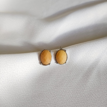 Par de brincos estilo botão oval em tom bege com lapidação facetada e base metálica, sobre tecido branco acetinado.