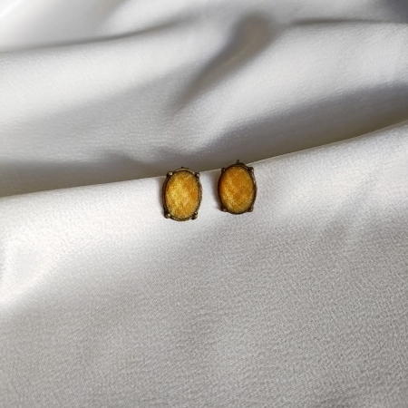 Par de brincos estilo botão oval em tom bege com lapidação facetada e base metálica, sobre tecido branco acetinado.