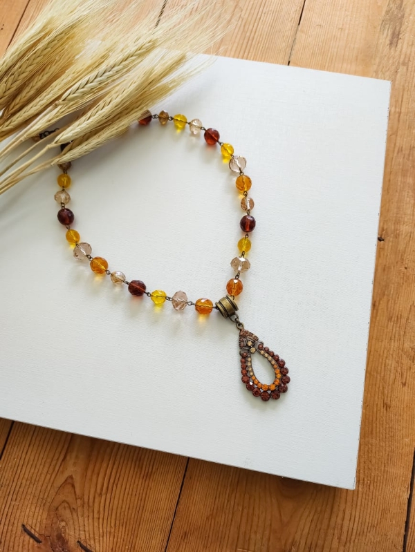 Colar boho com cristais em tons de âmbar e caramelo, com pingente vintage oval e fundo branco decorado com ramo de trigo.