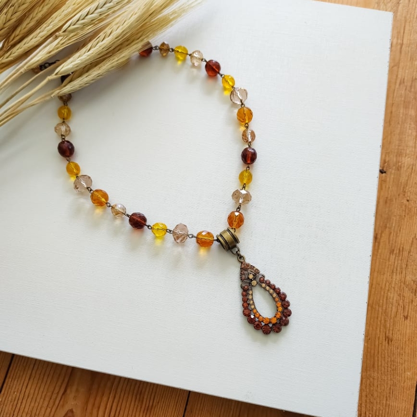 Colar boho com cristais em tons de âmbar e caramelo, com pingente vintage oval e fundo branco decorado com ramo de trigo.