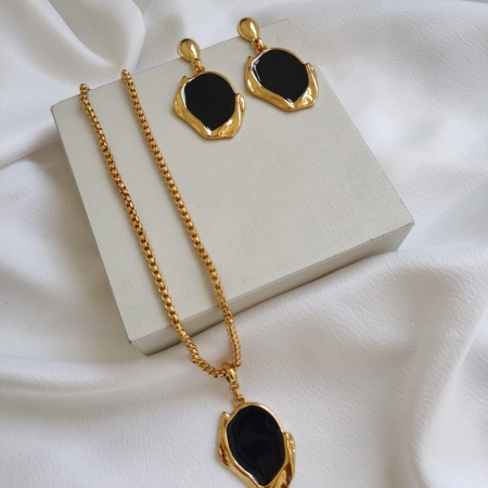 Conjunto de colar e brincos folheados a ouro com pedra preta, dispostos sobre tecido branco.