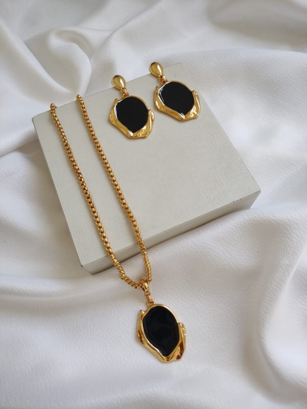 Conjunto de colar e brincos folheados a ouro com pedra preta, dispostos sobre tecido branco.