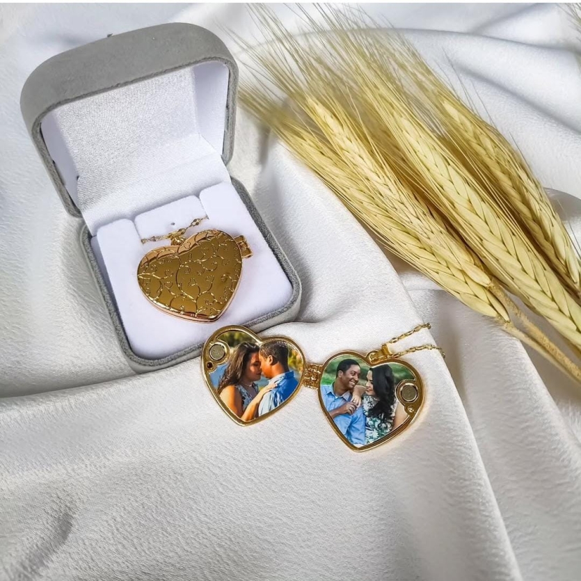 Relicário dourado em formato de coração com fotos personalizadas de casal em seu interior, acompanhado de caixa de veludo cinza e ramo de trigo sobre tecido branco.
