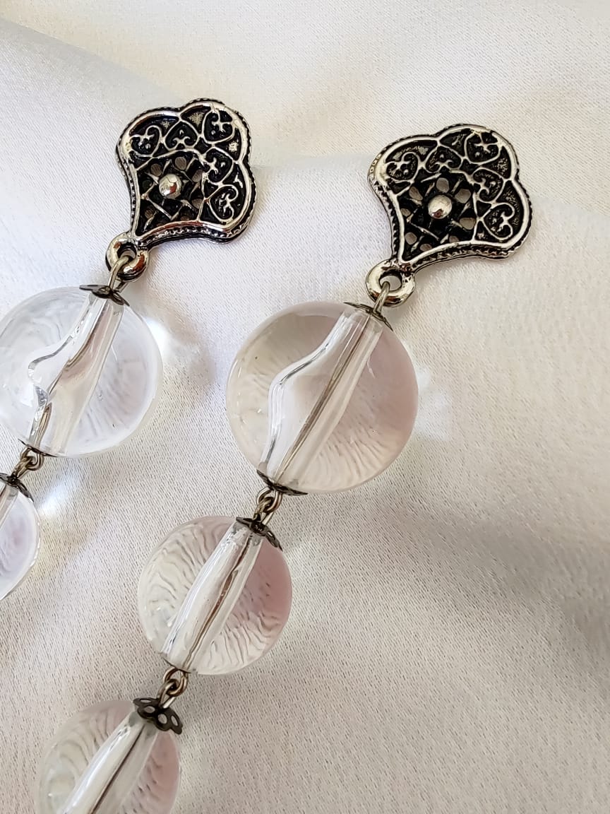 Brinco longo com contas transparentes lapidadas e base metálica trabalhada com detalhes vazados.